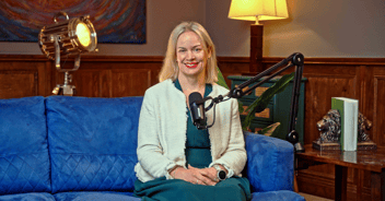 Image of a smiling person wearing a cream jacket and green dress, on a blue sofa. A podcast microphone is in front of her. 